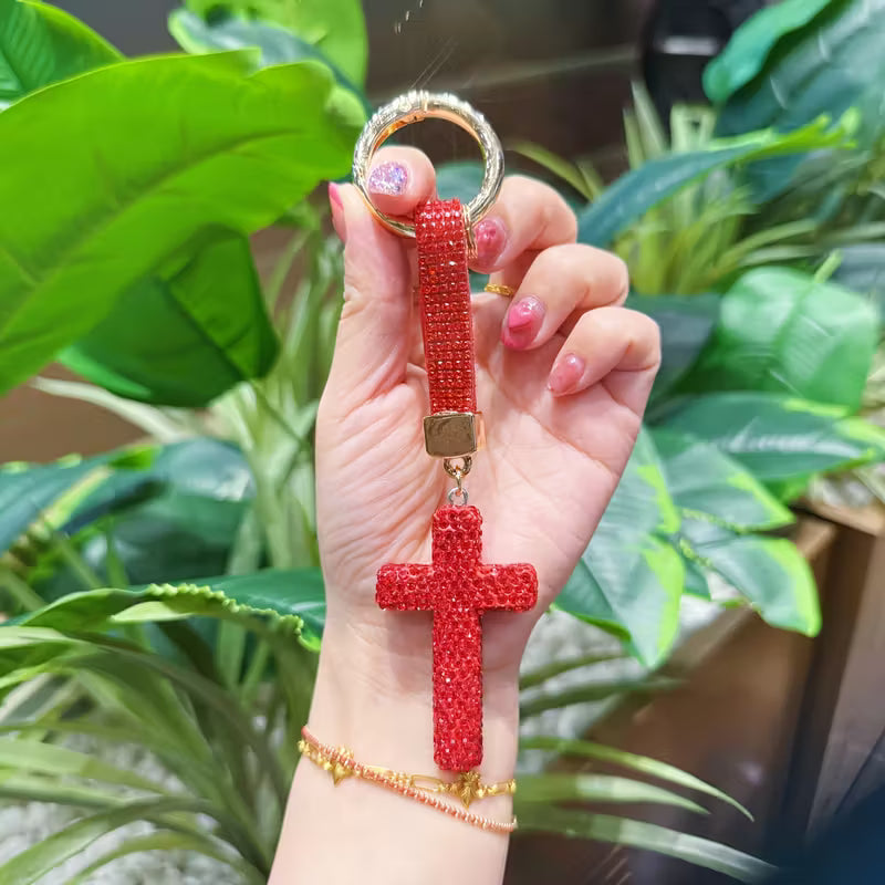 Hand holding a red cross keychain with green plants in the background