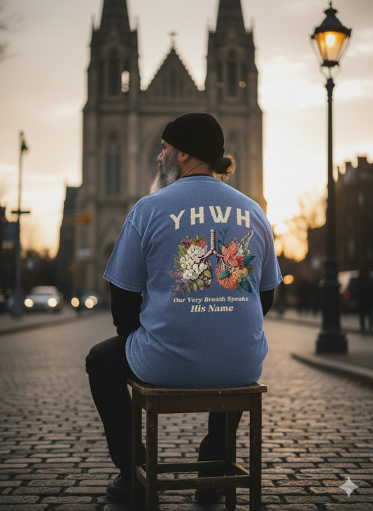 Person wearing a blue t-shirt with 'YHWH' and religious text, sitting on a stool in an urban setting.