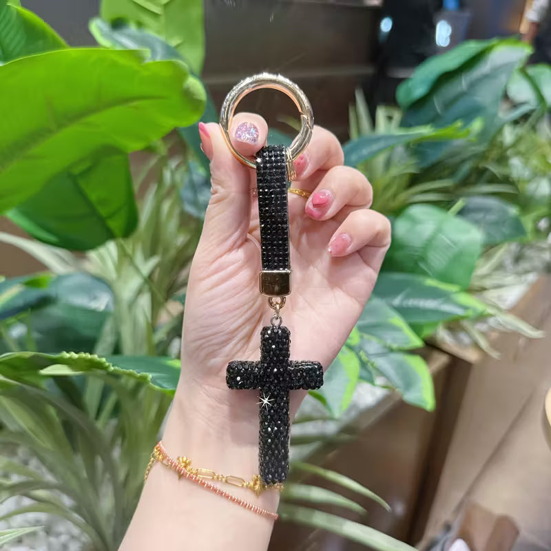 Hand holding a keychain with a cross design in front of green plants