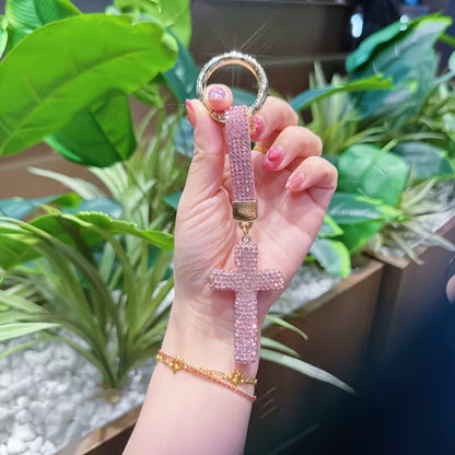 Hand holding a pink beaded cross keychain with plants in the background